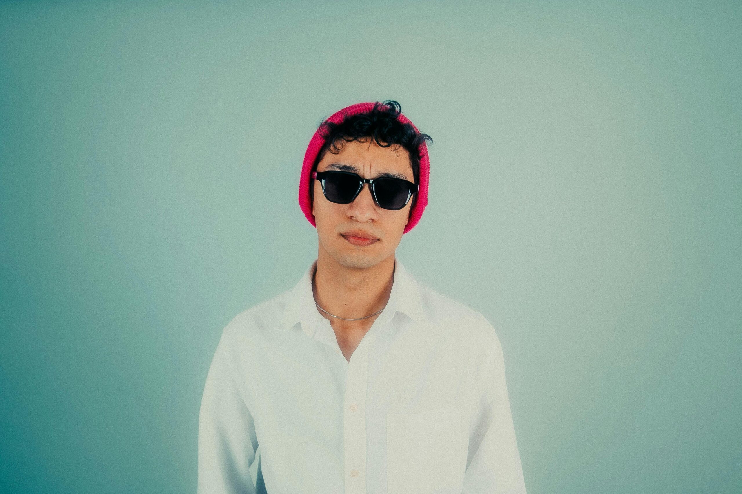 Fashionable young man wearing sunglasses and a pink beanie, set against a minimalist pastel background.